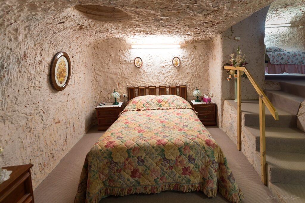 quarto subterrâneo em coober pedy, ao centro uma cama coberta por um acolchoado, luminárias na parede, escada para a superficie na lateral direita, móvel à esquerda no canto inferior da foto, um quadro em formato circular na parede esquerda.