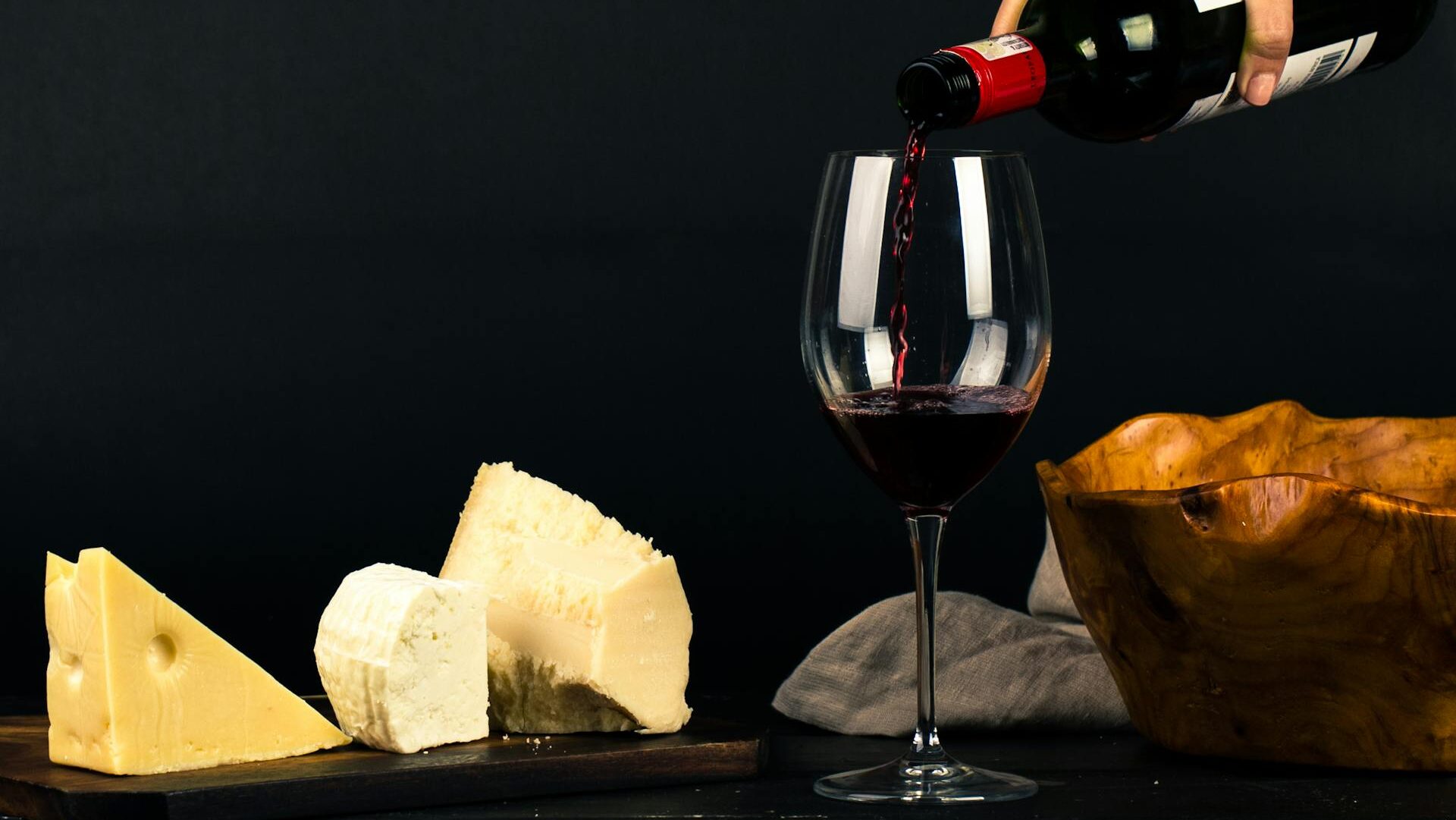 garrafa de vinho servindo uma taça, colocada sobre uma mesa onde se encontram queijos para harmonizar com a bebida