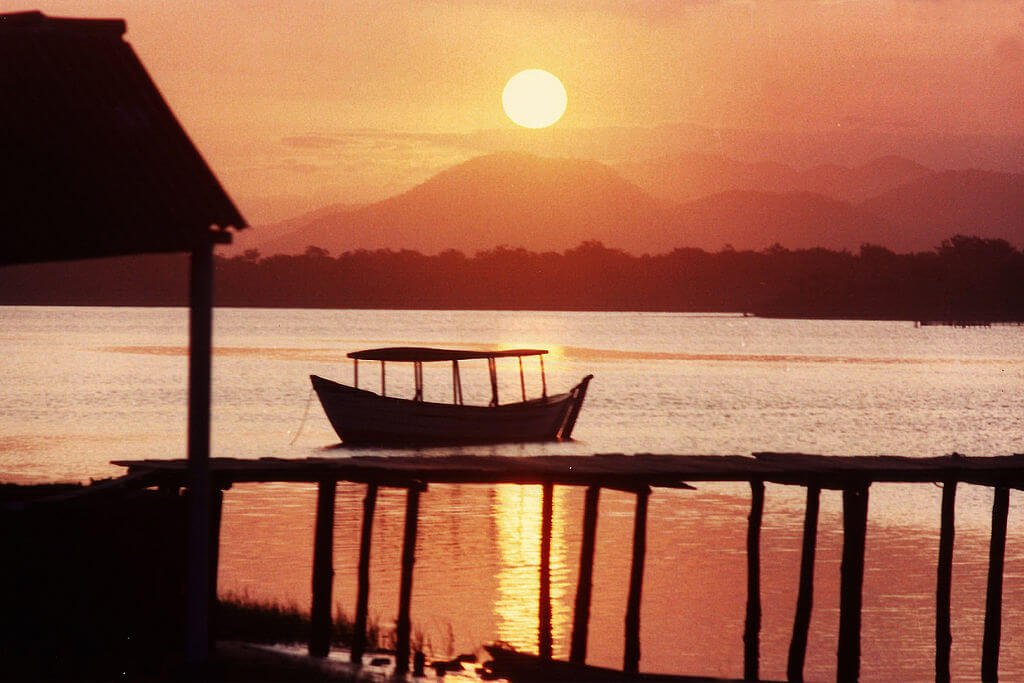 Fotos de viagens são ótimas para recordação. Esta foto está mostrando por do sol, tonalidades alaranjadas, sol acima das montanhas, silhueta da vegetação, trapiche na parte inferior da foto, à esquerda cobertura de uma barraca.