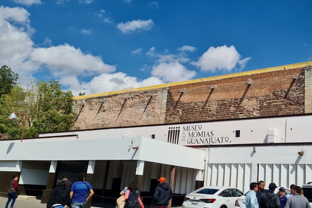 fachada externa do museu das misteriosas múmias de guanajuato, com grupo de visitantes reunidos ao lado externo do museu. dia de ceu azul com algumas nuvens. uma copa de árvore ao lado esquerdo da foto.