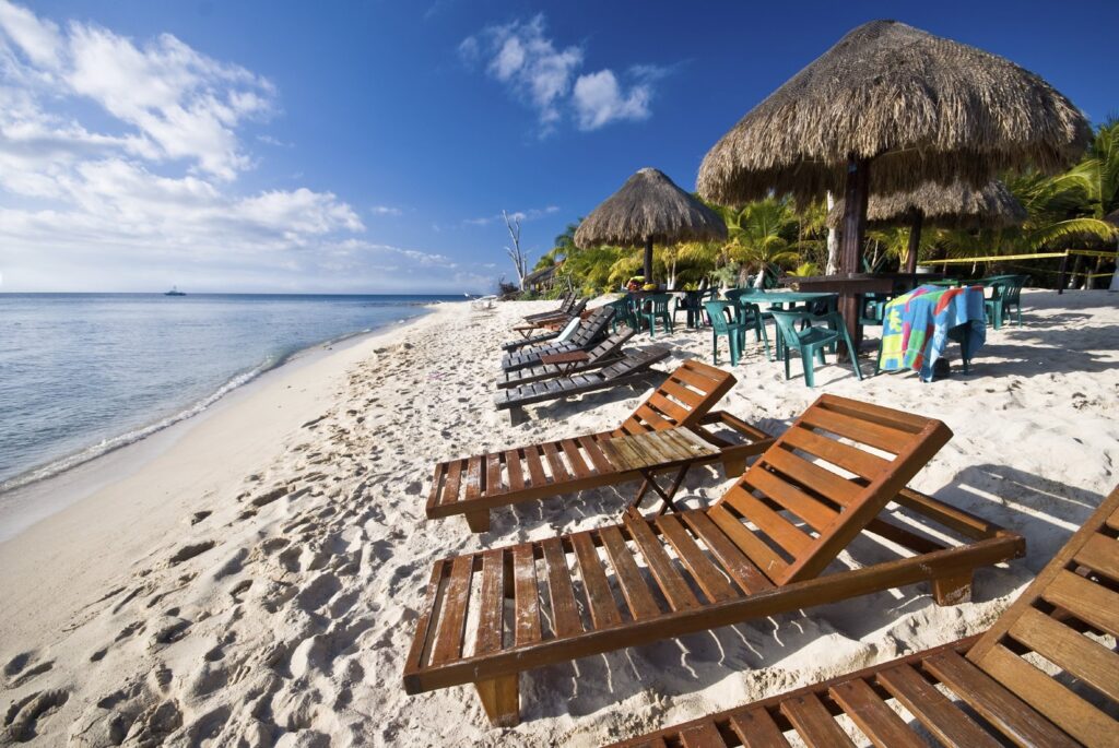 Praia Cozumel, Riviera Maya, um ótimo destino para viagens com crianças, amplas cadeiras de madeira sobre beira da praia com areia branca, água do mar à esquerda, à direita quiosques, na parte superior da foto, céu com poucas nuvens.