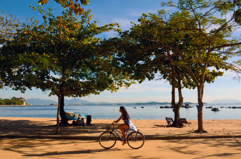 Mulher andando de bicicleta no acostamento junto à praia de Paquetá, no Rio de Janeiro. Duas árvores na calçada da orla e o mar ao fundo. Céu azul parcialmente nublado.