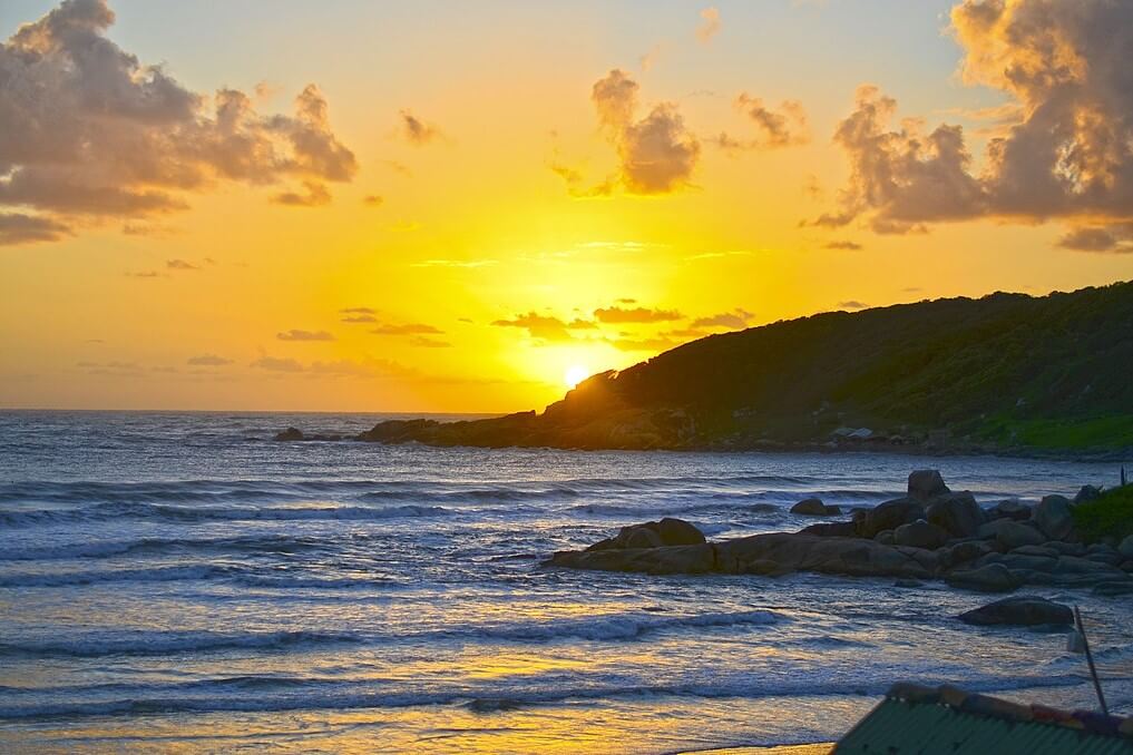 Nascer do sol na praia do Rosa, céu com tons de alaranjado com poucas nuvens, silhueta de formação geológica (morro) à direita, e outros rochedos na praia, mais próximos de onde foi registrada a foto. Praia do Rosa é um Tesouro Entre as Praias de Santa Catarina.