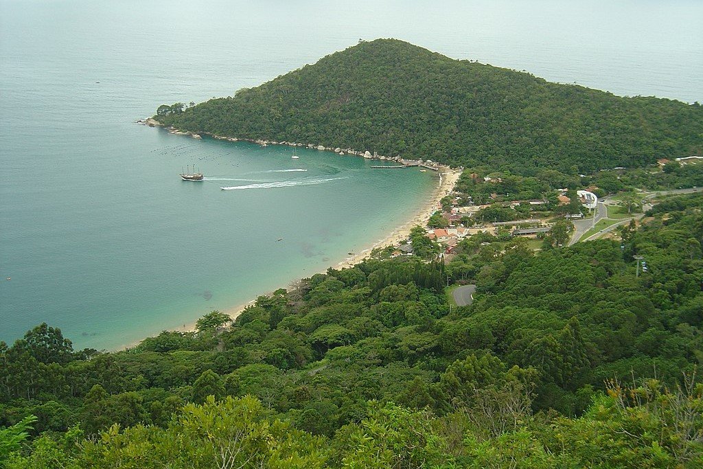 Enseada em Balneário Camboriu. Vegetação litorânea típica, embarcação de passeio no mar. Águas das praias, ao fundo oceano Atlântico.