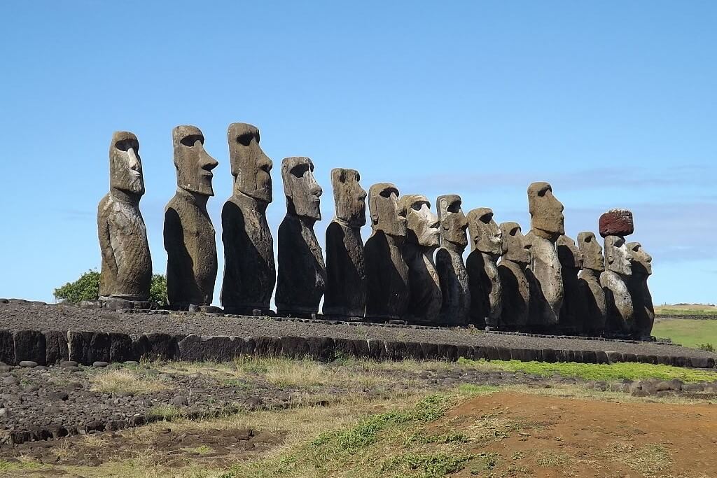 Quinze estátuas Moais na Ilha de Páscoa com os rostos virados para a direita da foto, sobre uma estrutura de suporte, em frente terreno sem cobertura vegetal, céu completamente azul, pouquíssimas e quase imperceptíveis nuvens ao longe.