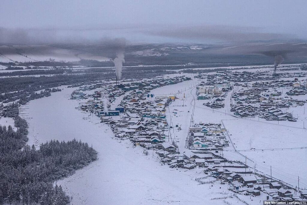 Vista aerea de Verkhoyansk mostrando inúmeras casas e o solo cobertos por neve, passando uma noção de frio extremo que as pessoas enfrentam. Vegetação típica desta região ao lado esquerdo da foto.