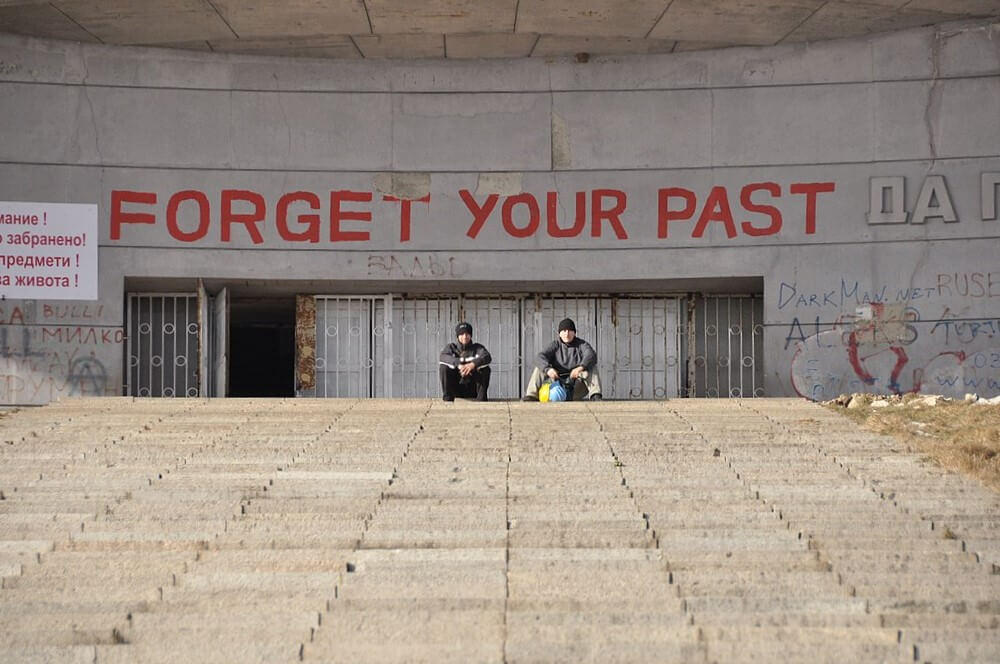 Escadaria que conduz ao pavimento onde se encontra acesso para o monumento de Buzludzha. Dois jovens sentados junto o acesso ao monumento. Acima, pixação no Monumento com a mensagem em inglês 