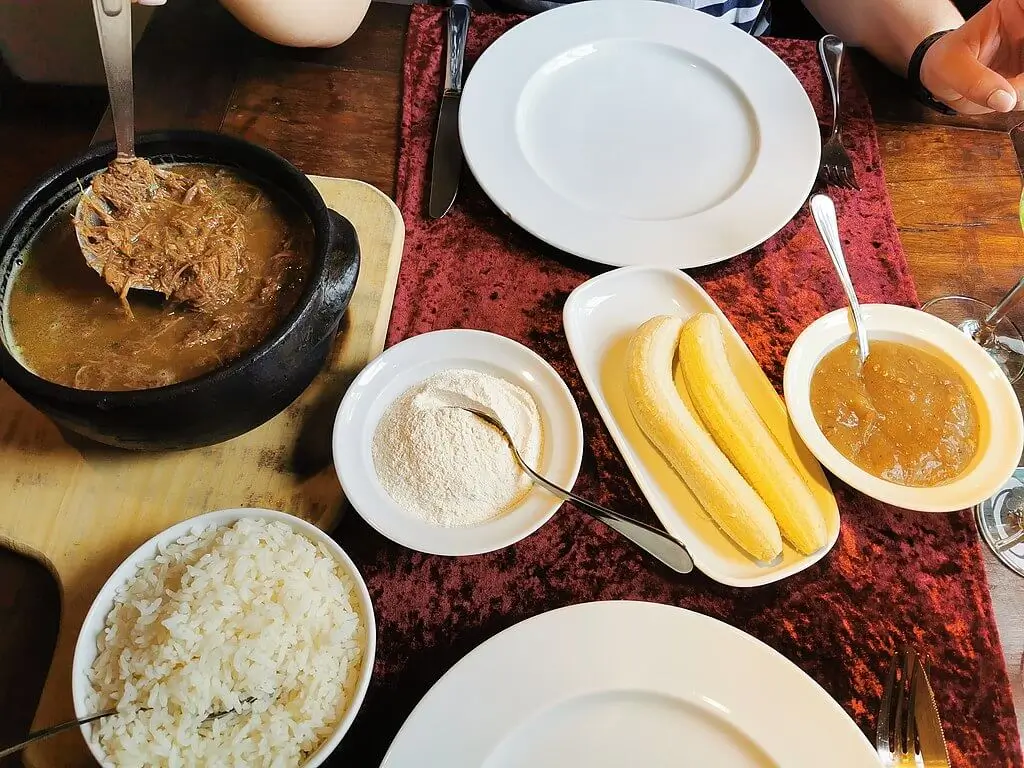 Panela de ferro à esquerda com uma porção de barreado, 6 outros pratos, sendo um com arroz branco, outro com duas bananas, um com algum tipo de creme ou molho e dois pratos vazios. mesa de madeira rústica com uma toalha avermelhada servindo de base para os dois pratos das pessoas que se alimentarão do barreado. Tábua de madeira servindo de base para a panela de ferro. Barreado é um dos ícones quando se fala em sabores rurais brasileiros