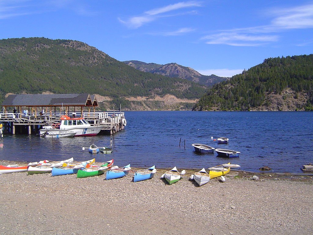Margem do Lago Lagar constituído por seixos e sedimentos de menor dimensão, caiaques de diferentes cores, lancha, pier, as águas do lago Lacar, montanhas cobertas por vegetação ao fundo. Considerada a irmã menor de Bariloche, San Martín de los Andes, na Argentina, é uma excelente opção para ir além de Bariloche.