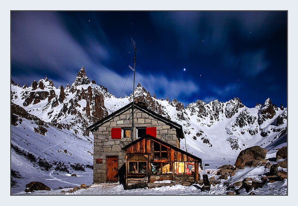 Chalé de madeira, iluminado no interior, em frente à construção de pedras, com aberturas vermelhas. Ao fundo montanhas da Cordilheira dos Andes, Cerro Catedral é um dos tesouros da, agora, Bariloche Revelada.