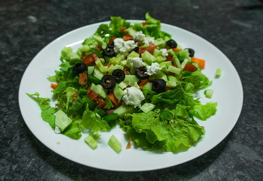 Mesa com um prato branco acima, servindo salada mediterrânea (pepino em cubos, tomates, cenouras, pimentões e azeitonas, coberta com queijo feta e servida sobre uma cama de alface). A iguaria é uma das 11 receitas fáceis com limão Siciliano.